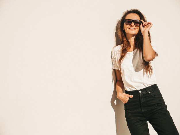 Портрет красивой улыбающейся модели в солнечных очках. Девушка одета в летнюю хипстерскую белую футболку и джинсы. Модная девушка позирует возле стены на улице