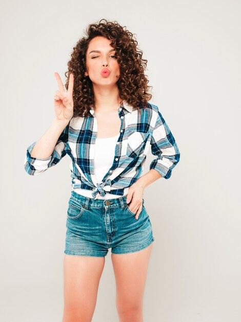 Portrait of beautiful smiling model with afro curls hairstyle dressed in summer hipster clothes.Sexy carefree girl posing in studio on gray background.Trendy funny and positive woman shows peace sign