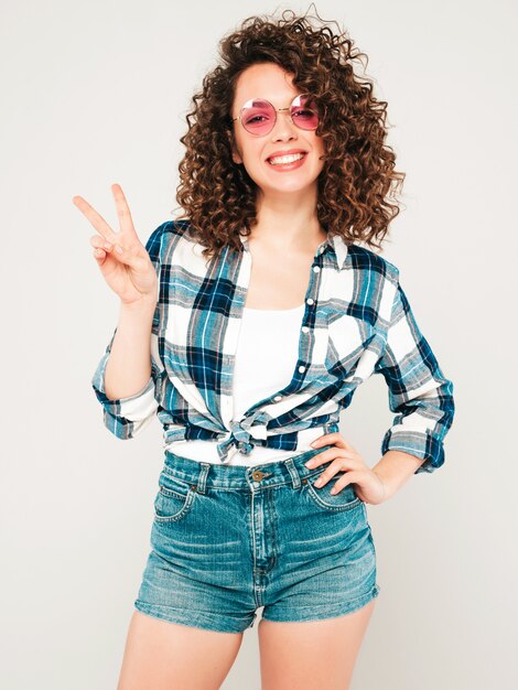 Portrait of beautiful smiling model with afro curls hairstyle dressed in summer hipster clothes.Sexy carefree girl posing in studio on gray background.Trendy funny and positive woman.Shows peace sign