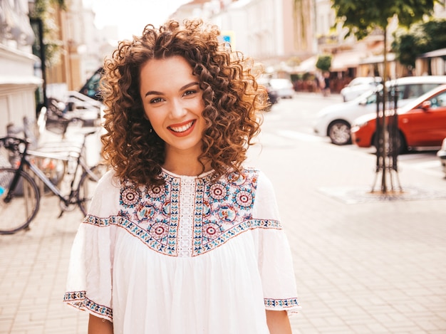 Portrait of beautiful smiling model with afro curls hairstyle dressed in summer hipster white dress.