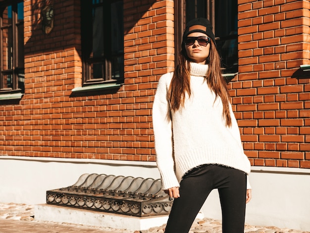 Portrait of beautiful smiling model. Woman dressed in warm hipster white sweater and beanie. She posing in the street