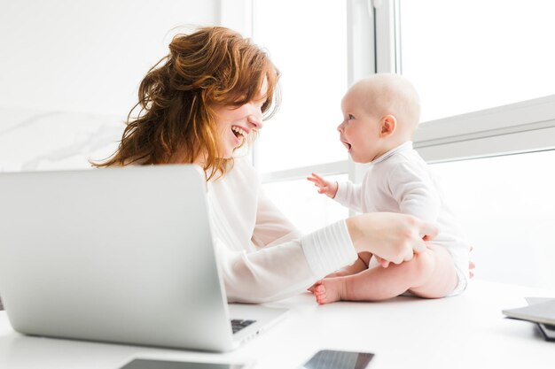 랩톱에서 작업하는 동안 앉아 있고 그녀의 귀여운 작은 아기를 행복하게 보고 있는 아름다운 미소 어머니의 초상화