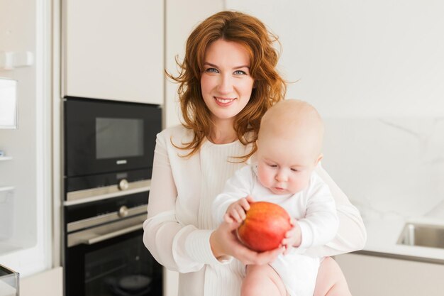 Portrait of beautiful smiling mother standing on kitchen and happily looking in camera while holding her cute little baby and big red apple in hands