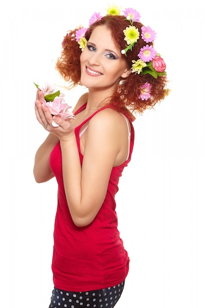 Portrait of beautiful smiling redhead ginger woman in red cloth with yellow pink colorful flowers in hair isolated on white holding flowers
