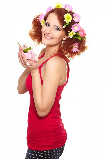 Portrait of beautiful smiling redhead ginger woman in red cloth with yellow pink colorful flowers in hair isolated on white holding flowers