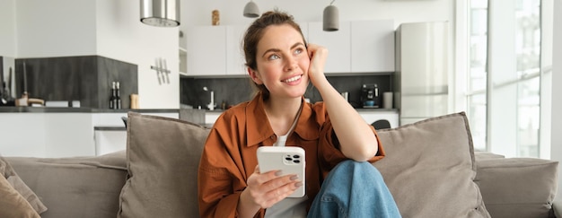 Портрет красивой улыбающейся женщины со смартфоном, сидящей дома на диване, думающей и смотрящей