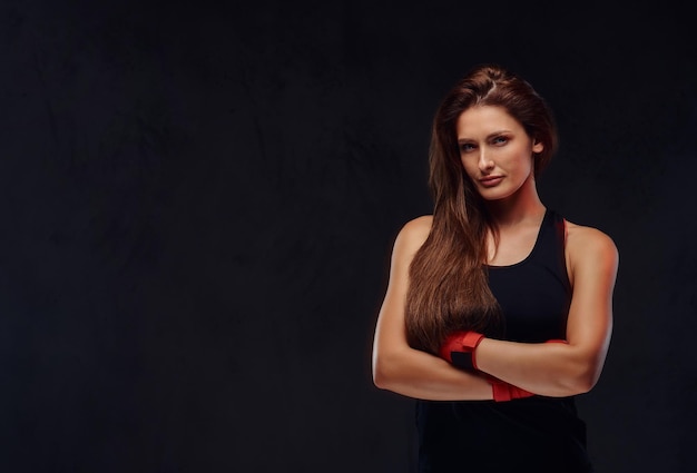 Portrait of a beautiful sportive female boxer in sportswear and bandaged hands, posing with crossed arms. Isolated on a dark textured background.