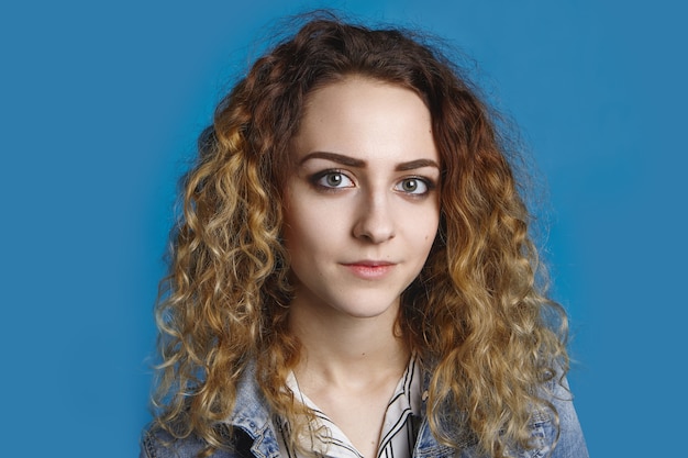 Portrait of beautiful student girl with clean perfect skin and wavy light hair posing , dressed in denim jacket against blank blue wall wall with copy space for your advertising content