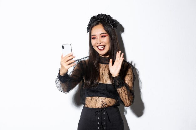 Portrait of beautiful and stylish asian woman in gothic lace dress saying hi, waving hand at smartphone camera