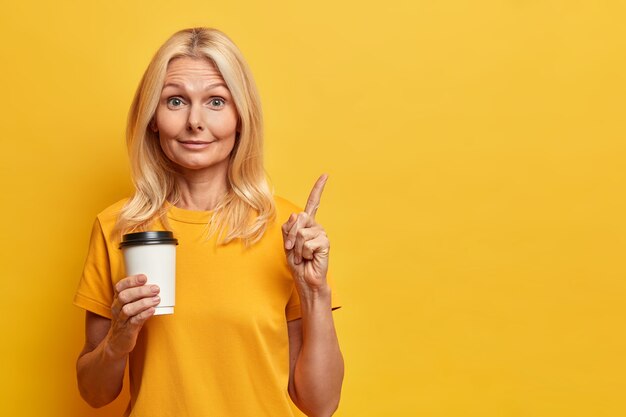Portrait of beautiful surprised woman points at upper right corner holds take away coffee gets idea during break has healthy skin and blonde hair 