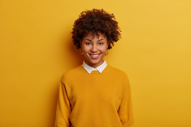 Portrait of beautiful tender woman has cheerful smile, wears casual jumper with white collar, stands against vivid yellow space, looks directly at camera