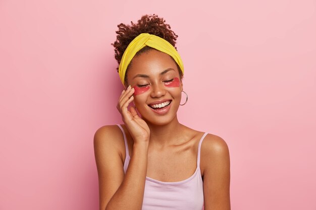 Portrait of beautiful woman carries eye patches, enjoys effect of perfect skin, keeps eyes closed, wears yellow headband and casual t shirt