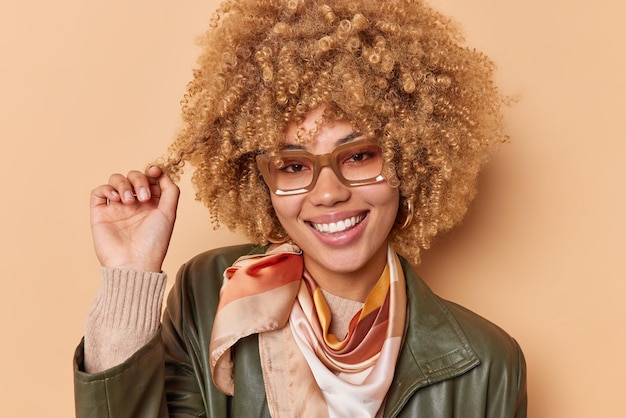 Portrait of beautiful woman curls hair smiles broadly shows white teeth being in good mood wears spectacles and stylish clothes isolated over beige studio wall has pleased facial expression.