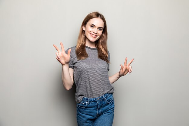 Portrait of beautiful woman dressed in casual clothes smiling at camera while showing peace sign with fingers isolated over beige wall in studio