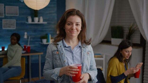 Portrait of beautiful woman holding beer glass looking into camera during night party in background ...