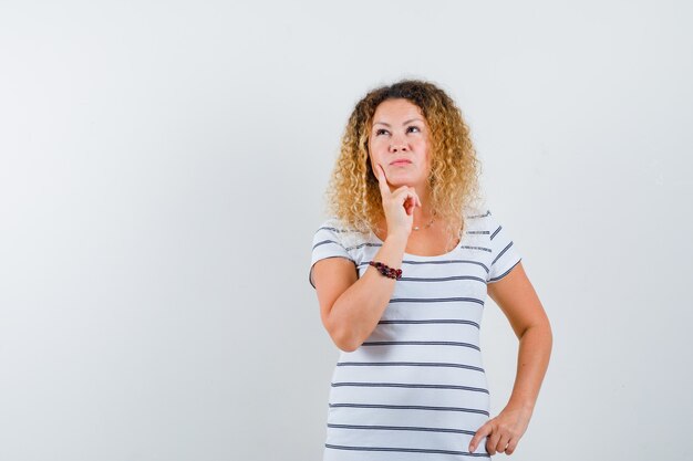 Portrait of beautiful woman holding finger on cheek, looking up in t-shirt and looking thoughtful front view