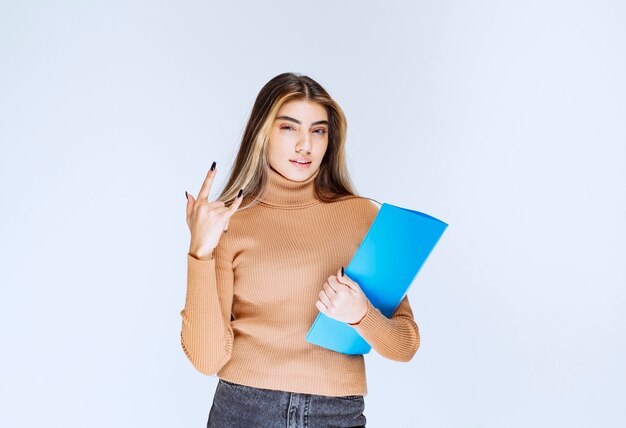 Portrait of a beautiful woman holding a folder and showing rock sign against white wall .