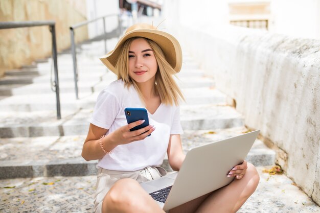 Portrait of beautiful woman holding mobile phone and laptop sitting on the summer city stairs