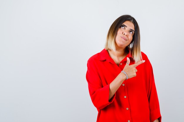 Portrait of beautiful woman pointing right, looking up in red blouse and looking thoughtful front view