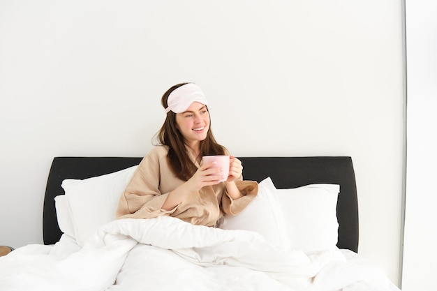 Free photo portrait of beautiful woman in sleeping bag and pyjamas drinking coffee in bed enjoying her bright