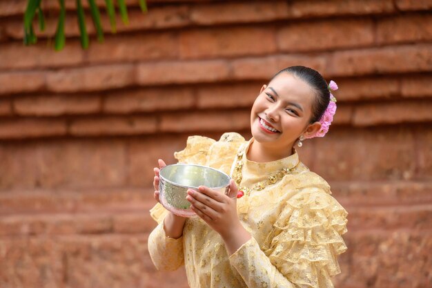 水ボウルを保持している寺院でタイの伝統的な衣装とソンクラン祭りの肖像画の美しい女性と水祭りでタイ文化を笑顔