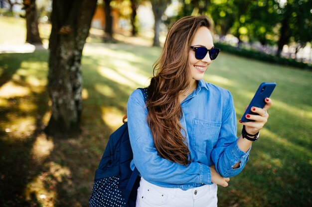 Portrait of a beautiful woman in sunglasses typing on the smart phone in a park with a green unfocused background