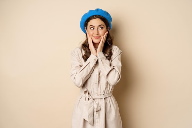portrait of beautiful woman in trendy outfit, touching her face and looking dreamy, yearning smth, standing against beige background. people concept