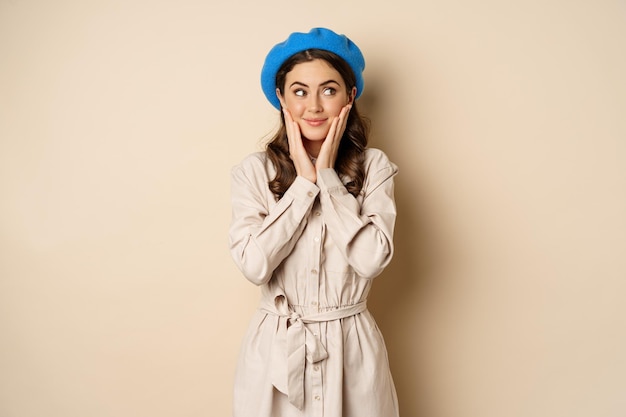 Free photo portrait of beautiful woman in trendy outfit, touching her face and looking dreamy, yearning smth, standing against beige background. people concept