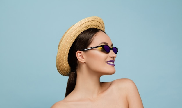 Portrait of beautiful woman with bright make-up, hat and sunglasses on blue studio