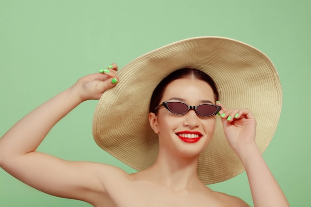 Free photo portrait of beautiful woman with bright make-up, hat and sunglasses on green studio background. stylish and fashionable make and hairstyle. colors of summer. beauty, fashion and ad concept. smiling.