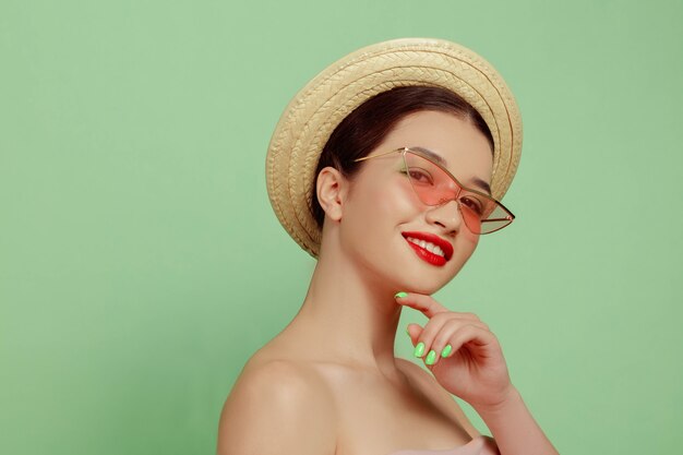 Portrait of beautiful woman with bright make-up, hat and sunglasses on green studio background. Stylish and fashionable make and hairstyle. Colors of summer. Beauty, fashion and ad concept. Smiling.