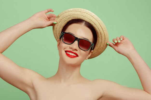 Портрет красивой женщины с ярким макияжем, шляпой и солнцезащитными очками на зеленой студии