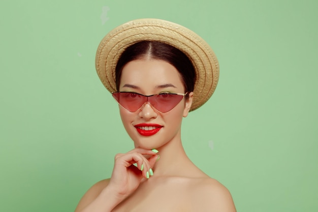 밝은 메이크업, 빨간 안경 및 녹색 스튜디오 배경에 모자와 아름 다운 여자의 초상화. 세련되고 세련된 메이크업과 헤어 스타일. 여름의 색상. 미용, 패션 및 광고 개념. 포즈.