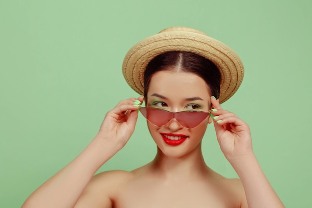 밝은 메이크업, 빨간 안경 및 녹색 스튜디오에 모자와 아름 다운 여자의 초상화