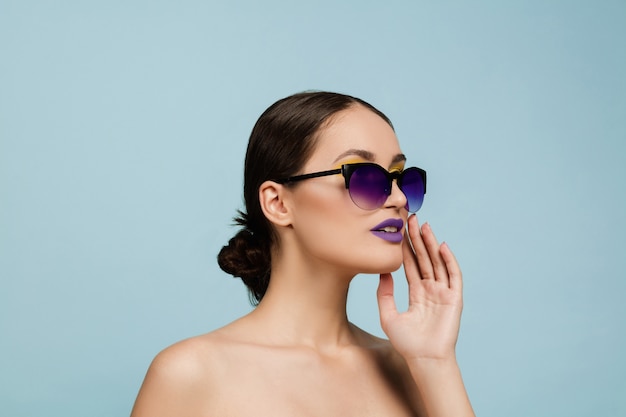 Free photo portrait of beautiful woman with bright make-up and sunglasses on blue studio background. stylish, fashionable make and hairstyle. colors of summer. beauty, fashion and ad concept. calling somebody.