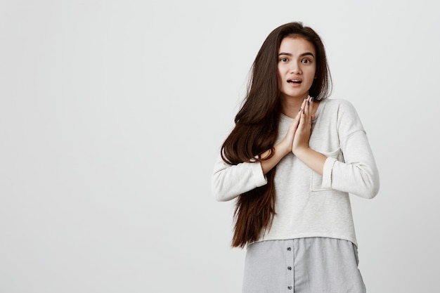 Portrait of beautiful woman with long dark hair dressed casually having hopeful and excited expression keeping her palms together in amazement. Happy brunette emotional female glad to receive present.