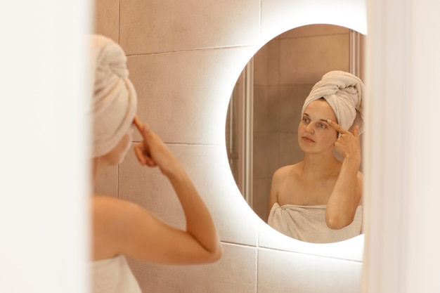 Portrait of a beautiful young adult woman with a towel on the head standing in the bathroom and examining her face in the mirror, touching her eyebrow.