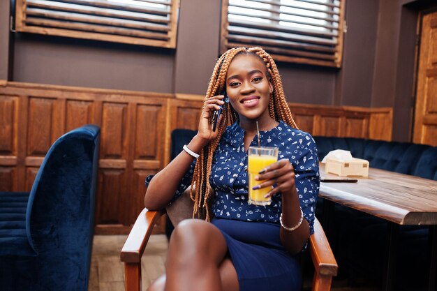 Portrait of beautiful young african business woman wear on blue blouse and skirt sitting with juice in cafe and speak on phone