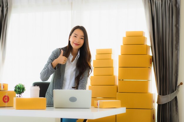 肖像画の美しい若いアジアビジネス女性自宅でラップトップ携帯電話で段ボール箱を出荷の準備ができていると仕事