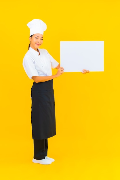 Foto gratuita ritratto bella giovane donna chef asiatica con cartellone vuoto bianco su sfondo giallo isolato