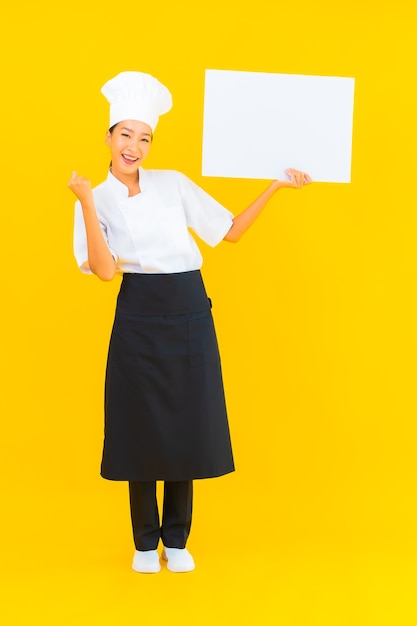 Ritratto bella giovane donna chef asiatica con cartellone vuoto bianco su sfondo giallo isolato