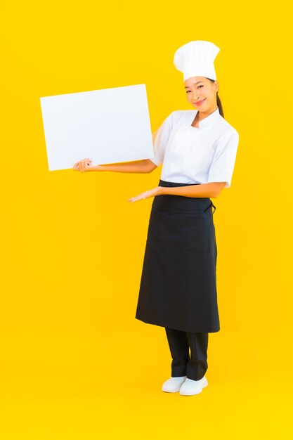 노란색 외진 배경에 흰색 빈 광고판이 있는 아름다운 젊은 아시아 요리사 여성 초상화