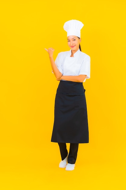 Женщина портрета красивая молодая азиатская в форме шеф-повара или повара с шляпой на желтом изолированном фоне