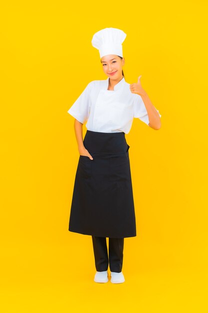 Женщина портрета красивая молодая азиатская в форме шеф-повара или повара с шляпой на желтом изолированном фоне