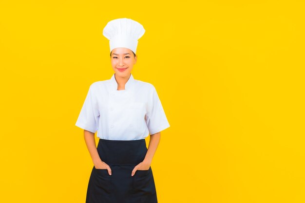 노란색 외진 배경에 모자를 쓴 요리사 또는 요리사 유니폼을 입은 아름다운 젊은 아시아 여성의 초상화