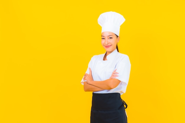 Ritratto bella giovane donna asiatica in chef o cuoco uniforme con cappello su sfondo giallo isolato