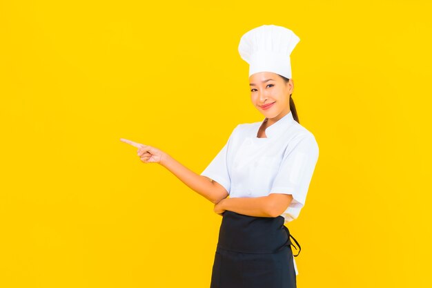 Ritratto bella giovane donna asiatica in chef o cuoco uniforme con cappello su sfondo giallo isolato