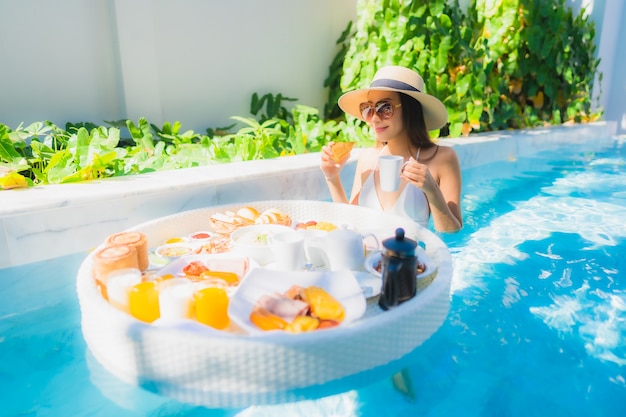 Улыбка красивой молодой азиатской женщины портрета счастливая с плавая завтраком в подносе на бассейне