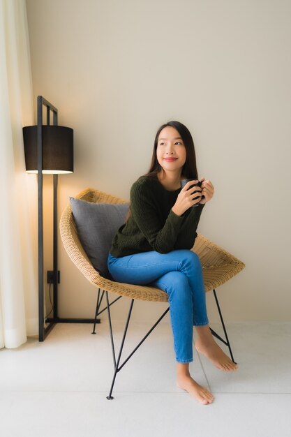 Чашка кофе владением женщины портрета красивая молодая азиатская и сидеть на стуле софы