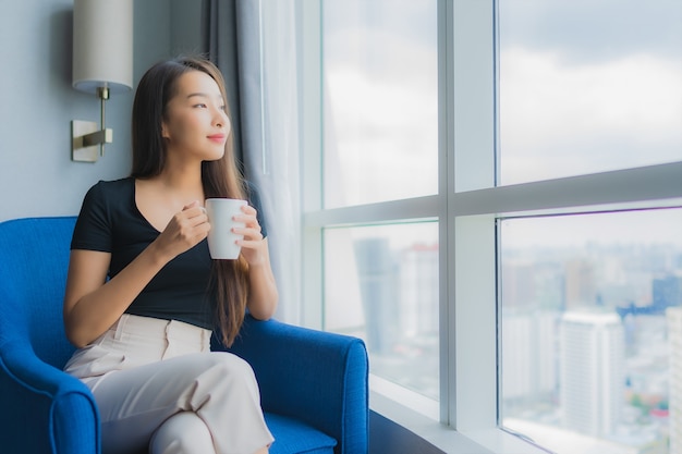 Чашка кофе владением женщины портрета красивая молодая азиатская на стуле софы в зоне живущей комнаты